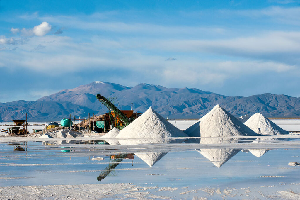 Industrial brine mining operation in a salt flat, with large white salt or mineral mounds reflected in shallow water. Mining equipment and tanks are visible in the background, with a backdrop of blue mountains under a partly cloudy sky.