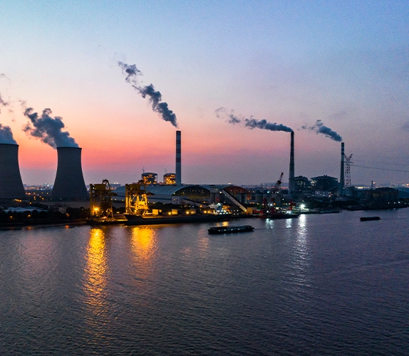 Coal-fired power plant at dusk with smokestacks emitting steam or smoke over a river, illuminated by industrial lighting.