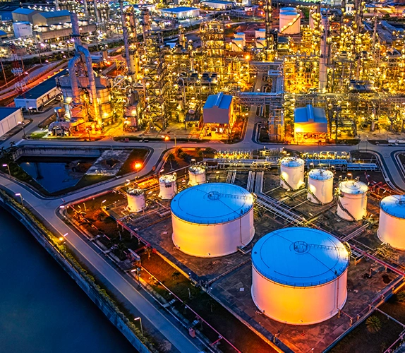 Aerial view of an illuminated oil and gas refinery at night, featuring large storage tanks, processing units, and surrounding waterways.